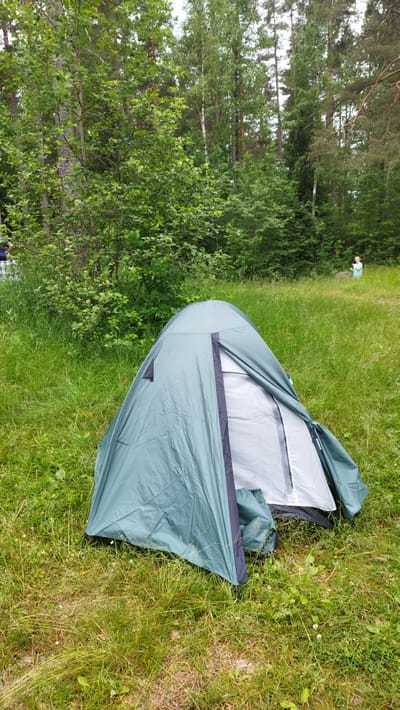 A solitary, partially open, teal-colored tent is pitched in a grassy clearing, nestled at the edge of a dense forest. The tent's entrance is unzipped, revealing a light-colored interior. Tall trees, with a mix of deciduous and coniferous species, form a thick backdrop, their green foliage illuminated by what appears to be diffused daylight, suggesting an overcast or cloudy sky. The ground is covered in lush, green grass, interspersed with smaller plants and some fallen leaves. In the background, further into the clearing and near the treeline, a small figure, possibly a child wearing a light blue shirt, stands next to a large rock, suggesting a moment of peaceful outdoor activity. The overall scene evokes a sense of nature, camping, and quiet solitude.