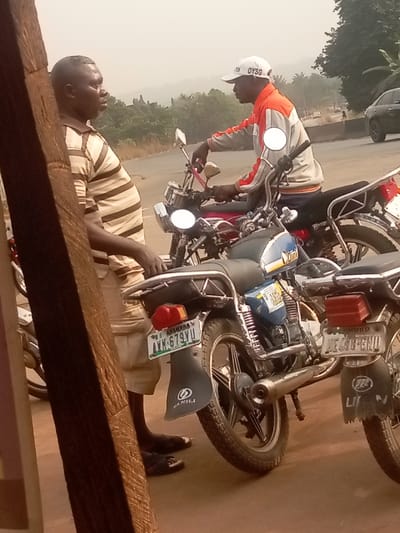 Two men converse near parked motorcycles. One man wears a striped shirt and beige shorts, standing near a wooden post. The other sits on a red motorcycle, sporting a white cap labeled "OYSG" and an orange-and-gray jacket. The motorcycles, one blue and one with a visible license plate "LEE-488-ENU", line the roadside. The background shows a road, other vehicles, and distant trees under a hazy sky.