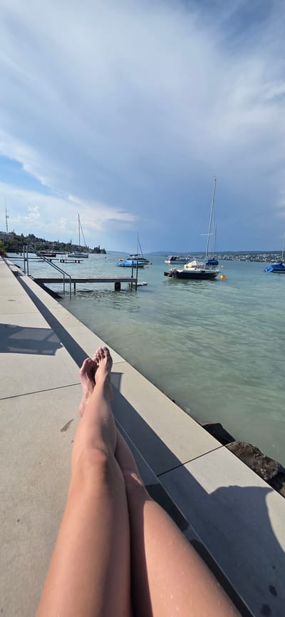 In Oberrieden, Switzerland, a person's legs and feet are in the foreground, resting on a concrete walkway next to a body of water. The person's legs are crossed, and their feet are bare. Water droplets are visible on their skin, suggesting they may have recently been in the water. The walkway runs alongside a lake, with several sailboats moored in the distance. The sky is partly cloudy, with a mix of blue sky and darker, dramatic clouds. Rocky elements are visible along the edge of the water.