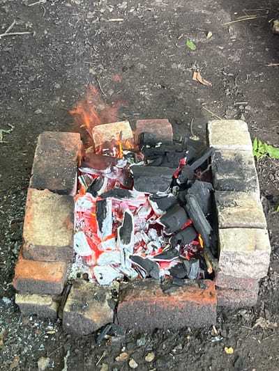 An outdoor scene depicts a rectangular pit constructed from weathered bricks, containing glowing red embers and black charcoal. Small flames flicker among the charcoal pieces. The ground surrounding the pit is composed of dark soil with scattered debris, including leaves and small twigs. The lighting suggests it is daytime. This scene is located in the city of Myrne, Ukraine. There are no people or animals present.