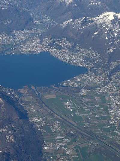 This is an expansive aerial photograph capturing a diverse landscape composed of towering mountains, a large lake, and significant urban and agricultural development.

The upper and right portions of the image are dominated by rugged mountain ranges. Many of the higher peaks and slopes are partially covered in white snow, suggesting either winter or early spring. The mountainsides display a mix of dark, forested areas, barren rock, and some lighter, possibly less vegetated patches. Valleys wind through these mountains, with roads and scattered settlements visible along their contours.

A large, deep blue lake occupies the central-left and middle sections of the image. Its shores are irregular, conforming to the mountainous terrain, and appear calm. Dense urban development, characterized by numerous buildings, can be seen clustered along the northern (left) and eastern (top-right) shores of the lake, forming what appear to be continuous towns or cities. Several marinas or dock areas are visible along the waterfront.

Towards the bottom-middle of the image, the lake narrows and transitions into a river system or a delta, leading to a broad, relatively flat plain. This plain is extensively cultivated, featuring a patchwork of rectangular agricultural fields. Some fields exhibit lighter rectangular structures, possibly greenhouses, indicating intensive farming. A distinct, straight canal or river runs through these agricultural areas, roughly parallel to a prominent road or highway. A bridge crosses this canal in the mid-ground.

Interspersed within the agricultural landscape and along the roads are further scattered residential areas and industrial complexes. In the lower-middle part of the image, there's a notable complex of buildings with adjacent cleared land that resembles a small airfield or industrial park. On the lower-left side, a large cluster of buildings, possibly an extensive fort or institutional complex, is visible nestled on a forested mountainside.

The image is taken during daylight hours, under clear skies, as indicated by the distinct shadows and bright illumination. The overall scene depicts a well-developed region with significant human activity, set against a dramatic natural backdrop. No text is visible in the image.