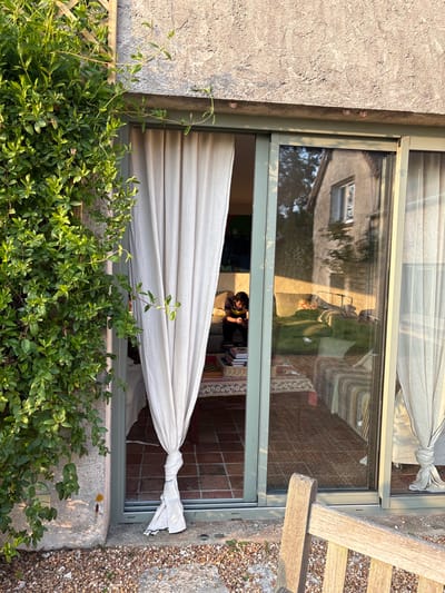 The scene is located in the city of Bû, France. An exterior vantage point captures a sliding glass door system leading into a residential interior. To the left, a concrete wall is covered by green climbing ivy. The ground is gravel, with a wooden bench partially visible in the lower right foreground. The gray-framed sliding door on the left is slightly open, revealing a light-colored curtain tied centrally. Through the open door and the transparent glass of the closed right door, an interior room with terracotta tile flooring is visible. One individual, with dark hair and wearing a dark top, sits on a light-colored sofa, looking down at an object in their hands. A low table in front of the sofa holds stacked books and a chessboard on a patterned cloth. The closed right door reflects an adjacent building, likely a two-story house with a visible window, under natural light, indicating an outdoor reflection.