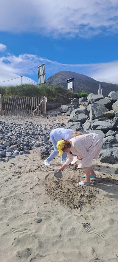 Two individuals are observed on a sandy beach in Pier Road, Ireland, with a large rocky outcrop and a grassy hill with a mountain in the background. The person in the foreground, wearing a pink patterned dress and blue sandals, is bent over, holding a grey stone in their right hand and appears to be placing it into a pile of sand. To the left, another person, dressed in a light blue top and white sneakers, is also bent over, facing away from the camera. The beach is covered in sand and small pebbles, with footprints visible in the sand. A wooden fence runs along the edge of the grassy area. The sky is blue with scattered white clouds.