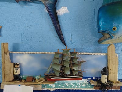 A wall decoration featuring a model ship is affixed to a blue, textured wall. The decoration includes two mounted fish specimens: a long, dark blue fish tail and a blue-green fish head. The model ship, centrally located, has dark sails and a hull with red, black, and white stripes. The ship rests on a platform painted with waves. To the left of the ship is a wooden carving of a sailor, while to the right is a miniature lighthouse. The decoration is framed by rope and pieces of wood. The setting is Cutler Bay, United States of America.