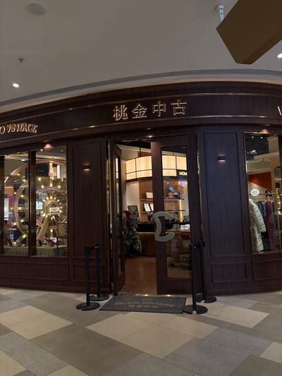 The image shows the exterior of a shop called "MOMO VINTAGE" located in Heping District, China. The shop has a dark wood facade with gold lettering. The entrance is open, with a welcome mat that reads "MOMO VINTAGE" and Chinese characters. Inside the shop, a Christmas tree is visible. There are also display windows showcasing merchandise, including a large spinning wheel with lights and what appears to be handbags and clothing. Two stanchions are placed outside the entrance. The floor in front of the shop is tiled in a geometric pattern of beige, gray, and taupe. The overall impression is of a retail establishment in a commercial area.