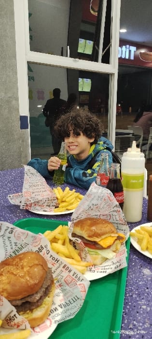 A young boy with curly hair sits beside a window, holding a green beverage. The table in front of him has two plates with burgers and french fries. Also visible on the table are two bottles of Coca-Cola and a container of sauce. The entire scene is captured at an angle, showing a glimpse of the restaurant's interior reflected in the window.