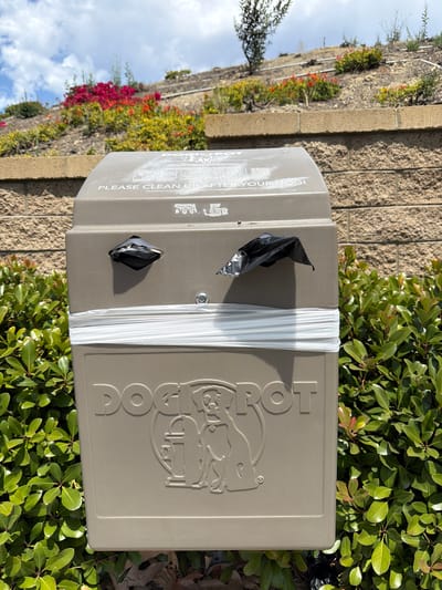 A dog waste station stands in front of a low stone wall, with a slope of flowering bushes and trees behind it, under a cloudy sky. The station is a tan, rectangular box with a domed top. The top of the box has text that reads "PLEASE CLEAN UP AFTER YOUR DOG!". There are two black plastic bags protruding from openings on the front of the station. A large roll of white plastic bags is wrapped around the middle of the station. The front of the station also features the embossed logo "DOGIPOT" with a silhouette of a dog and a trash can. The station is surrounded by green bushes. The background shows a textured brick wall and a raised embankment with landscaping. The location is in Irvine, United States of America.