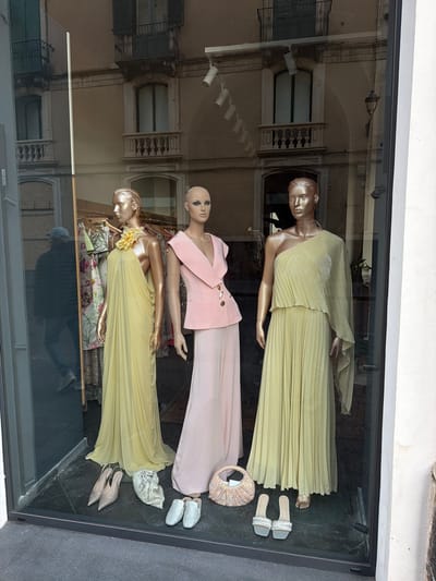 The image shows a window display of a clothing store, likely in Acireale, Italy, based on the reflection of buildings with architectural features typical of the region. Three mannequins are dressed in formal wear. The top mannequin wears a pleated yellow dress, accompanied by a pair of silver and white sandals and a decorative handbag. The middle mannequin is dressed in a pale pink dress with a matching blazer featuring decorative buttons. The bottom mannequin is wearing a pale yellow flowing gown with a large yellow flower embellishment, paired with taupe high heels and a beaded bag. The window reflection shows buildings with arched windows, balconies, and street lighting. The display is set against a dark gray floor.