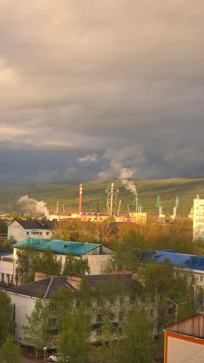 The image depicts a cityscape in Lensk, Russia. A large industrial complex with a tall, red-striped chimney stack and several cranes is visible amidst a dense green forest. Smoke or steam plumes rise from the industrial area. Adjacent to the forest and industrial zone are several multi-story residential buildings with various colored roofs (blue, grey, brown), partially obscured by trees with green foliage. The sky shows a division between a cloudy, grey area and a lighter, potentially sunlit, section.