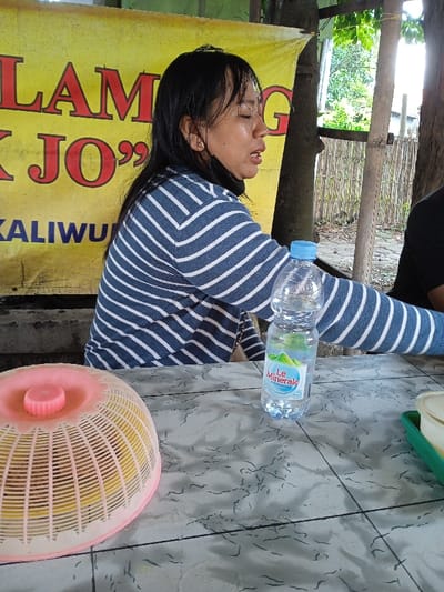 A woman in a blue and white striped sweater sits at a table with a bottle of Le Minerale water. A food cover with a pink lid and a green tray are on the table. A yellow sign with red text hangs behind her. Part of a person in a black shirt is also visible on the right. The setting appears to be an outdoor dining area, with wooden posts and a fence in the background.