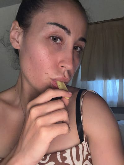 A close-up, low-angle shot captures a woman in Dromolaxia - Meneou Municipality, Cyprus, with her face partially visible as she eats a piece of food, possibly a French fry, which she holds between her fingers and brings to her mouth. Her head is tilted, and her gaze is directed downwards, away from the camera. She has her hair pulled back. Her skin shows some texture and what appear to be blemishes. She is wearing a dress with a tan and white patterned design, and a black bra strap is visible. The background consists of sheer curtains and indistinct shadows.