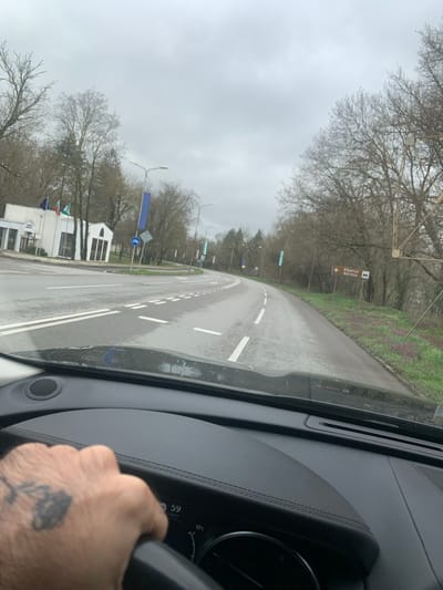 The image depicts a view from inside a car driving on a wet road in Ushintsi, Bulgaria. The road has white lane markings, and there is a bicycle lane sign visible on the left side of the road. Several buildings and trees are also visible along the roadside. The sky is overcast. A hand is visible on the steering wheel of the car. The dashboard indicates a temperature of 10°C and a speed of 59 km/h. A sign indicating the location of "Абритус" (Abrittus) is visible.