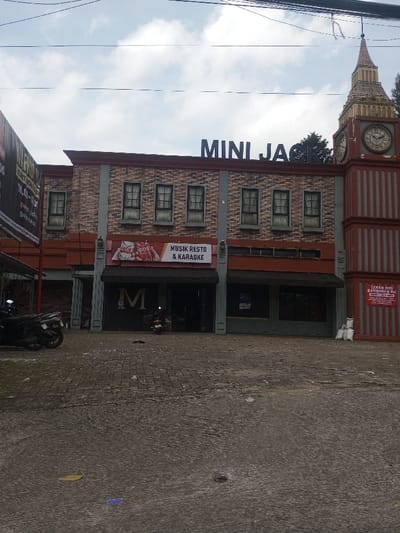 The image shows a building labeled "MINI JACK." The building has a brick-like facade with multiple windows on the second story and a large "M" sign above the entrance. To the right, there is a tower resembling Big Ben. The sign above the entrance says "MUSIK RESTO & KARAOKE". Two motorcycles are parked in front of the building. The foreground is a paved area with some debris. There's a sign in Indonesian on the right side of the building. The sky is overcast.