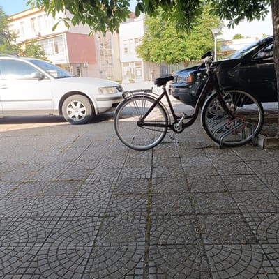 This image depicts a street scene in Lyubimets, Bulgaria, characterized by a tiled pedestrian area in the foreground and cars and buildings in the background. A dark-colored SUV is parked to the right, partially obscuring a burgundy bicycle. The bicycle is positioned in a bike rack, with its front wheel locked in place. To the left of the bicycle, a white car is parked. In the background, buildings with a mix of architectural styles are visible, including some with balconies and air conditioning units. Green trees with dense foliage are present, casting shadows on the ground and partially obscuring the sky. The overall scene suggests an urban environment on a sunny day.