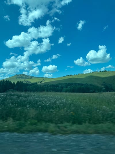 The image depicts a rural landscape with rolling green hills under a bright blue sky dotted with fluffy white clouds. The foreground shows a blurry field of green grass and wildflowers, suggesting movement, possibly from a vehicle. In the midground, a dense band of dark green trees forms a contrast against the lighter green of the hills. The hills themselves are covered in lush vegetation, with some areas appearing darker due to shadows. The overall impression is one of a serene, natural setting in a temperate climate, consistent with a location like Verkh-Aya, Russia, during daylight hours. There are no people or animals visible in the frame.