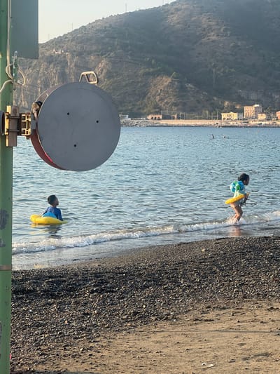 This image depicts a coastal scene on a sunny day in Sestri Levante, Italy. The specific location is consistent with a beach facing a promontory, likely the area around Sant'Anna or a beach in the eastern part of Sestri Levante, with the distinct coastal defenses and buildings visible in the distance.
In the left foreground, a large, grey, circular industrial reel, approximately 0.8 meters in diameter, is mounted on a vertical green metal pole with a brown bracket. Ropes or cables are partially visible attached to the pole.
The midground features the sea and a pebble beach. The water is calm, displaying light blue and green tones. Two children are present in the shallow water. To the left, a child, approximately 5-8 years old, wearing a blue long-sleeved top, is seated in a yellow inflatable swim float with a blue section, positioned with their back mostly to the camera, looking towards the distant coastline. To the right, a second child, approximately 4-7 years old, wearing light-colored swim bottoms and a multi-colored long-sleeved top, is actively moving through the breaking waves near the shoreline. This child wears yellow and blue/green inflatable arm rings and holds a small, dark object in their left hand. Small, dark specks, possibly distant swimmers or buoys, are visible further out in the water. The immediate shoreline is composed of dark, wet pebbles, transitioning to lighter, dryer sand further up the beach to the right.
The background is dominated by a large, rugged, and vegetated hillside or mountain, featuring exposed rock formations and sparse trees. Utility poles are visible on the upper slopes. At the base of the distant hill, a developed coastline is visible, including multiple multi-story buildings and a concrete breakwater or coastal defense structure with visible archways extending into the sea. The scene is illuminated by bright, natural daylight.