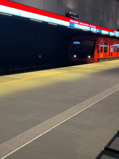 The image displays an underground metro station platform at Aalto University Metro Station in Espoo, Finland. A red metro train, unit 316-A, is partially visible on the right side of the frame, positioned alongside the platform. The train's front displays "TAPIOLA HAGALUND" as its destination, and its headlights are illuminated. The train carries the HSL (Helsinki Region Transport) logo. The platform is paved with grey square tiles and features a yellow safety line running along its edge closest to the tracks. A lighter-colored, textured band with a prominent white line traverses the platform floor in the foreground. Above the tracks, a large illuminated sign with a red background and white lettering reads "Aalto-yliopisto Aalto-universitetet Aalto University," accompanied by directional icons. The metro tunnel to the left of the train is dark, contrasting with the brightly lit platform and overhead signage. No individuals are present on the platform. Black metal grating is visible in the bottom right corner.