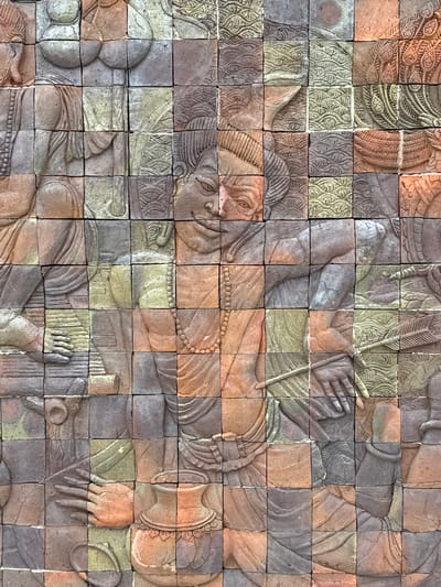 The image shows a detailed bas-relief sculpture on a brick wall, located in Chang Khoeng, Thailand. The sculpture depicts human figures in a narrative style, with each brick forming a tessellated part of the overall image. The figures appear to be engaged in various activities, with close-ups showing hands holding objects, draped clothing, and facial features. Decorative patterns of waves and floral motifs are integrated into the background of the relief. The color palette is dominated by earthy tones of terracotta, brown, grey, and muted green, suggesting natural materials and weathered texture. The composition suggests a historical or mythical scene, possibly from Thai folklore or religious art.