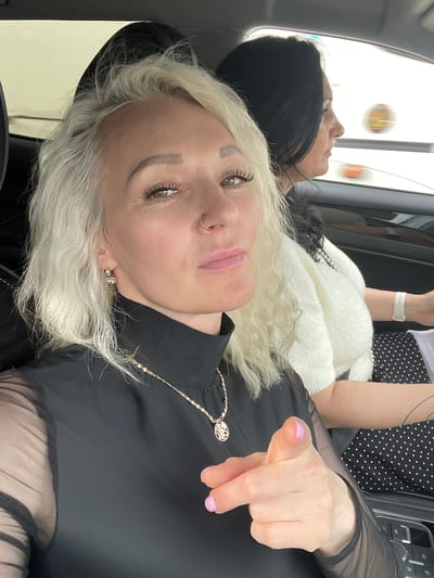 The image shows two women in a car. The woman in the foreground, with blonde hair and a black top with sheer sleeves, is looking at the camera and pointing her index finger forward. She is wearing a necklace with a circular pendant and a nose ring. Her nails are painted light purple. Behind her, a woman with dark hair is visible in the passenger seat, wearing a white fuzzy sweater. The car appears to be in motion, as indicated by the blurred background of what looks like a road and possibly other vehicles. The overall setting suggests they are traveling within or near the city of Yam-Izhora, Russia, given the metadata associated with the image.
