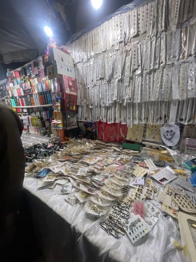The image shows a stall at an outdoor market, likely in Emure-Ekiti, Nigeria, at night. The stall is filled with an abundance of merchandise, primarily jewelry and perfumes. On the left, shelves are stacked with numerous perfume bottles of various brands and sizes, some in colorful packaging. Above the perfume shelves, more products are displayed, including what appear to be phone cases or similar small accessories.

The main focus of the stall is a long table covered with a white, shimmering fabric. This table is piled high with various types of jewelry, including necklaces, earrings, bracelets, and rings, most of which are packaged in plastic. There are also several racks of earrings on display. The background features a wall displaying numerous necklaces and chains, each individually packaged. A bright overhead light illuminates the scene. A person, partially visible on the far left, is facing away from the camera, suggesting they are a vendor or a customer. The overall impression is of a busy, well-stocked market stall operating after dark.