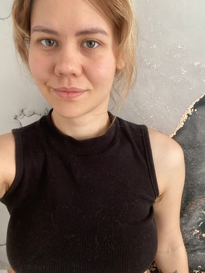 A selfie shows a woman with blonde hair and hazel eyes. She is wearing a black tank top. The background is a mottled gray and black wall, with gold flecks. The photo does not contain location information or objects related to the city of Dyadkovo, Russia.