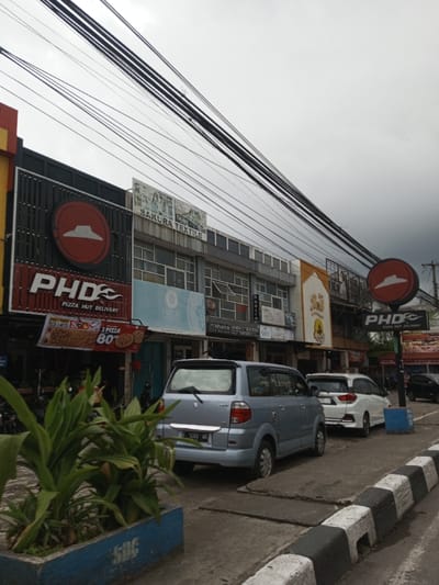 The image depicts a street view with several commercial buildings. Prominently featured are a PHD Pizza Hut Delivery restaurant, with signage advertising pizza deals, and a Sakura Textile shop. A light blue Suzuki minivan is parked in front, with a white Honda behind it. Power lines stretch across the sky above the buildings. A planter box with plants and "SOC" painted on the side is visible in the foreground, along with a black and white curb.