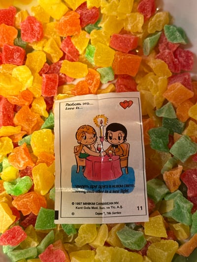 A scattered collection of colorful candied fruit pieces in shades of red, orange, yellow, and green fills the frame. Centered amongst the candy is a "Love is..." cartoon card. The cartoon depicts a couple at a candlelit table, with text in Russian and English: "Love is... seeing each other in a new light." Copyright information for MINIKIM CARIBBEAN NV is also printed on the card. The number "11" is visible in the upper right corner.
