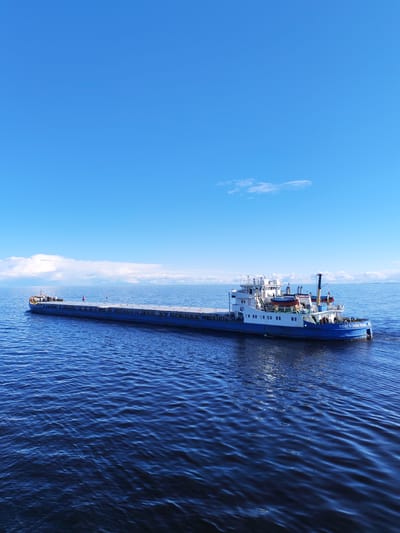 The image depicts a long, blue and white cargo barge sailing on a deep blue body of water under a bright blue sky with scattered white clouds. The barge has a multi-level white superstructure at its stern. The name "ВОЛГО-ДОН 5060" is painted on the stern of the ship in white letters. The image does not contain any visual information that would allow the determination of the specific location in Novaya Svobodka, Russia. The focus is solely on the barge on the water. There are no visible people or actions occurring.