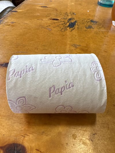 A roll of toilet paper with a floral pattern and the word "Papia" printed on it is placed on a wooden surface. The toilet paper is white with a light purple imprint. The wooden surface is textured and appears to be a table or countertop. There are some dark smudges and stains on the wood, suggesting it may be an older or well-used surface. A small portion of a blue glass object and a white piece of material are visible in the upper left corner of the image. The lighting suggests it is daytime, and there are no indications of specific weather conditions or emotions. The location context provided is посёлок опытного хозяйства «Ермолино», Russia.