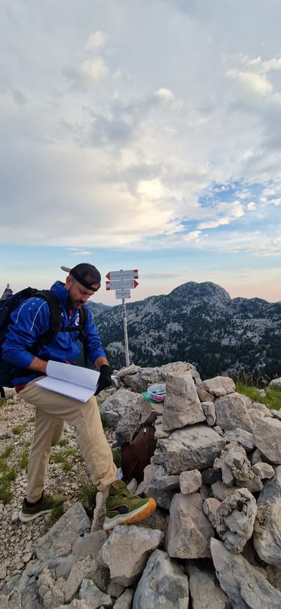 The image depicts a man standing on a rocky mountain summit in Gornje Polje, Montenegro, examining a notebook. He is wearing a blue jacket with white accents, beige pants, a black baseball cap, and black gloves. A backpack is visible on his left shoulder.

In the background, a mountain range covered with trees stretches across the landscape under a cloudy sky. A hiking signpost with red and white arrows is planted in the ground near the man. The foreground is dominated by a pile of large, grey rocks, with some green vegetation growing between them. A small plastic container filled with pens and pencils rests on the rocks to the man's right. A rusty metal object is partially visible among the rocks, behind the man's right leg. The lighting suggests it is either early morning or late afternoon.