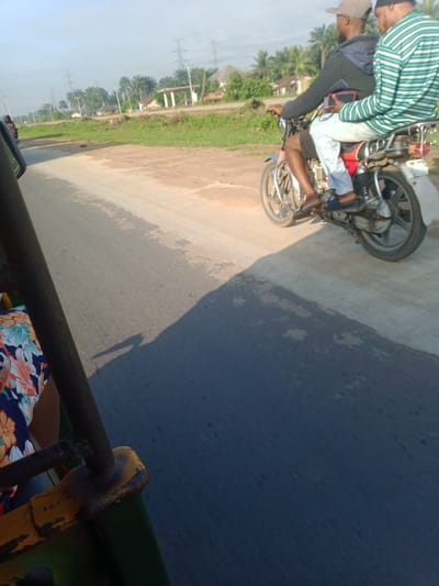 The image shows a scene in Asa Umu Nka, Nigeria. A motorcycle with two male passengers is moving on a paved road. The rider is wearing a grey cap, a black jacket, and light-colored jeans. The passenger wears a striped shirt, red cap, and light-colored jeans. The motorcycle is light in color with a red gas tank. A tricycle, from which the photo is taken, is partly visible in the foreground, with a floral pattern visible on the passenger's clothing. The environment includes greenery and utility poles in the background, with some buildings visible. The road is dark grey. The lighting suggests it is daytime.