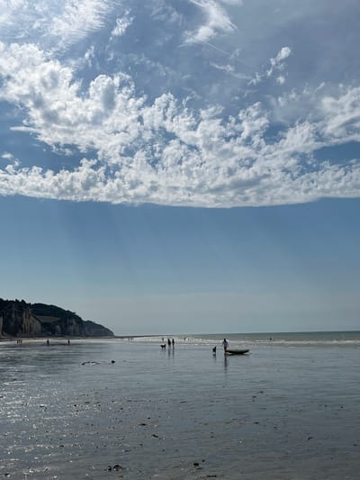 This media file depicts a serene beach scene at Hautot-sur-Mer, France. The image is divided into two distinct halves: the left side showcases a vast expanse of blue sky filled with dynamic, white cumulus clouds, some of which are elongated and streaky, hinting at movement and possibly the contrail of an aircraft. The right side of the image transitions to the beach and the sea.

The beach appears wet and reflective, with ripples and small puddles from the receding tide. Scattered across the sand are dark pebbles or shells, catching the light. In the distance, a gentle shoreline meets the calm sea, with the horizon line barely discernible. Along the water's edge, several figures are visible, appearing as small silhouettes against the bright, reflective surface. One group of people appears to be near two surfboards, suggesting a recreational activity. Further down the beach, more individuals are spread out, some walking, others perhaps observing the surroundings.

To the far right, a rugged cliff face rises from the edge of the beach, covered in green vegetation, providing a natural boundary to the scene. The lighting suggests it is daytime, likely late morning or early afternoon, given the bright sky and the reflections on the wet sand. The weather appears fair and calm, with no indication of strong winds or rain. The overall mood is one of tranquility and leisure, typical of a pleasant day at the seaside. There is no visible text in the image.