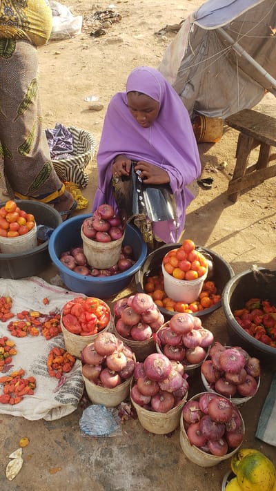 In Kawo, Nigeria, a woman wearing a purple hijab is sitting near an open-air market stall. She is positioned near buckets and baskets filled with produce including red onions, tomatoes, and peppers. An umbrella provides shade. Other individuals are partially visible in the foreground. The ground is sandy and shows signs of market activity.