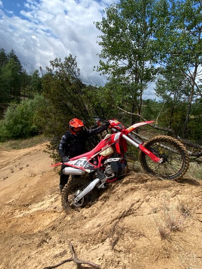 In the city of Парэцкі сельскі Савет, Belarus, a male motocross rider, wearing an orange helmet and black protective gear, is standing next to a red and white dirt bike. The motorcycle is partially submerged in muddy sand on a steep incline. The background consists of dense trees and a cloudy sky. The terrain is characterized by sandy mounds and vegetation, suggesting an outdoor off-road environment.