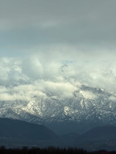 The image displays a wide-angle view of a mountainous landscape under an overcast sky, observed from the vicinity of Samtredia, Georgia. The upper third of the frame features a light blue-grey sky, partially covered by higher, thinner clouds. Below this, a dense formation of white-grey clouds partially obscures the upper and mid-sections of a prominent, snow-capped mountain range. The mountains show significant snow cover on their peaks and forested upper slopes, with dark tree clusters visible through the snow. Below this primary range, multiple strata of darker, forested hills recede towards the foreground, exhibiting a progressively muted blue-grey hue due to atmospheric perspective. Scattered light-toned specks, consistent with distant structures or dwellings, are faintly discernible on the lower slopes within the middle ground. The immediate foreground is marked by a dark, undifferentiated horizontal band, indicative of dense, distant vegetation or tree lines. No human figures or specific actions are discernible within the scene.