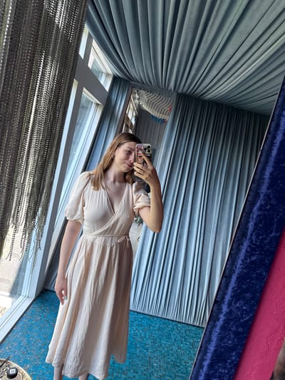 A woman is taking a selfie in a reflective space. She is wearing a light-colored, wrap-style dress that extends to her mid-calf. Her hair is light brown and styled in loose waves. She is holding a mobile phone in her left hand and is positioned in front of a window with strands of beaded curtains hanging in front of it.
The reflective space she is in appears to be a room or a display area, as suggested by the textured wall coverings in the background. A section of blue mosaic tiles is also visible, as are drapes that are blue and pink.
The setting seems interior and decorated. There is not sufficient information in the image to place the location in the city of Чёрное, Russia.