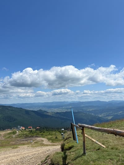 The image depicts a scenic mountain landscape in Chorna Tysa, Ukraine, under a bright blue sky with scattered white clouds. In the foreground, a wooden fence with rough-hewn logs runs diagonally across the frame, bordering a grassy slope. A dirt road curves uphill from the bottom left, leading towards a cluster of buildings nestled on the mountainside. These buildings appear to be part of a resort or village, with a few structures featuring red and white roofs. A ski lift, with chairs visible, ascends the slope in the middle ground, indicating a ski area. The surrounding mountains are covered in lush green forests, with rolling hills and valleys stretching into the distance. The overall scene suggests a serene and picturesque natural setting.