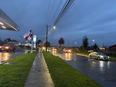 The image depicts a street scene in Osorno, Chile, under overcast skies at dusk or dawn. The ground is wet, suggesting recent rain. A concrete sidewalk runs alongside a grassy strip, bordering a street with visible vehicle traffic. A red pickup truck is turning into a gas station on the left, where the name "COPEC" is visible on a sign and flag. Other vehicles, including a silver sedan, are seen driving along the road. Power lines stretch across the upper part of the frame, and streetlights illuminate the scene. Buildings and trees line the street, adding to the urban setting.