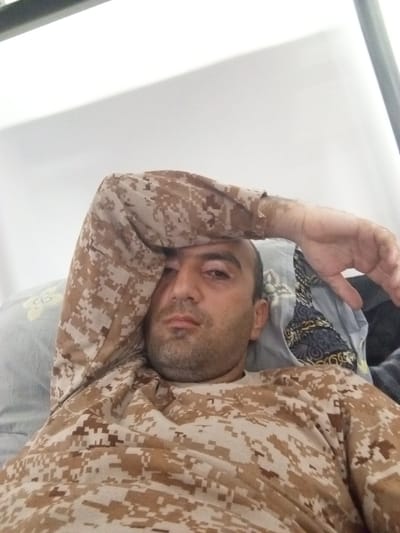 A man wearing a camouflage long-sleeved shirt is lying on a bed in what appears to be a room. His right arm is raised and resting on his forehead, and his head is slightly tilted. He has short, dark hair and stubble. Behind him, a pillow with a floral pattern is visible, as well as other bedding with a dark, patterned design. The background is a plain, light-colored wall, with some shadow cast on it. There is also a glimpse of a metal bar, possibly part of a bed frame, at the top of the image. The setting is indoors. The image does not contain any information that would allow for a specific location within Sarichashma Jamoat, Tajikistan.