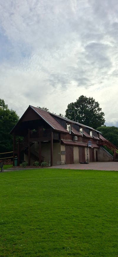 This image depicts a large, multi-story wooden building with a reddish-brown metal roof, situated in a grassy area with trees in the background. The building has a porch with stairs leading up to it, several windows, and two large doors at ground level. A basketball hoop and stand are visible in front of one of the doors. There are also air conditioning units mounted on the side of the building. The sky above is overcast with gray clouds. The scene appears to be an outdoor recreational area or a complex of buildings within a park or rural setting. The specific location in Valma, Estonia is not identifiable from the visual information alone, but the overall impression is of a functional building within a natural environment.