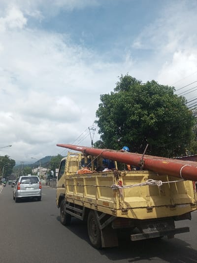 A yellow truck carries a long, red pipe secured with rope on a paved road. A silver car follows behind. Lush green trees and utility poles line the roadside under a bright sky with scattered clouds. The scene depicts everyday transportation and potential construction or infrastructure maintenance activities.