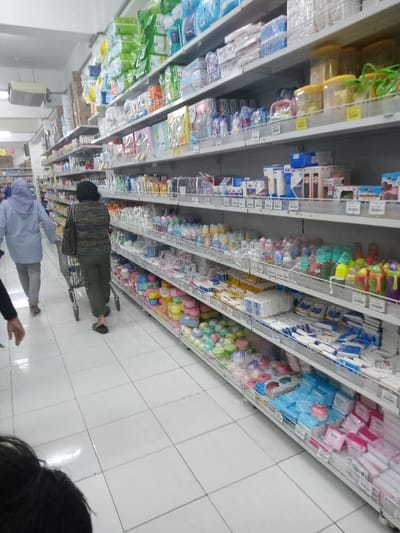 The image displays the interior of a brightly lit supermarket aisle in Nganjuk, Indonesia. The perspective looks down the length of the aisle, with multi-tiered metal shelving units lining the right side. These shelves are heavily stocked with various products, including numerous brands of diapers, baby bottles, pacifiers, baby wipes, small colorful plastic containers, and other personal care items, many with visible price labels.
Three individuals are present. In the lower-left foreground, the crown of a dark-haired person's head and a partial shoulder are visible. Approximately midway down the aisle, a person wearing a dark striped long-sleeved top, dark green cargo pants, and dark flip-flops walks away from the camera, pushing a silver shopping cart. Further ahead, a third individual, dressed in a light blue long-sleeved top, a light purple hijab, and grey pants, also walks away from the camera.
The floor is a clean, white tiled surface with dark grout lines. The scene is illuminated by overhead fluorescent lighting, and a white ventilation or air conditioning unit is visible mounted high on the left wall/ceiling junction.