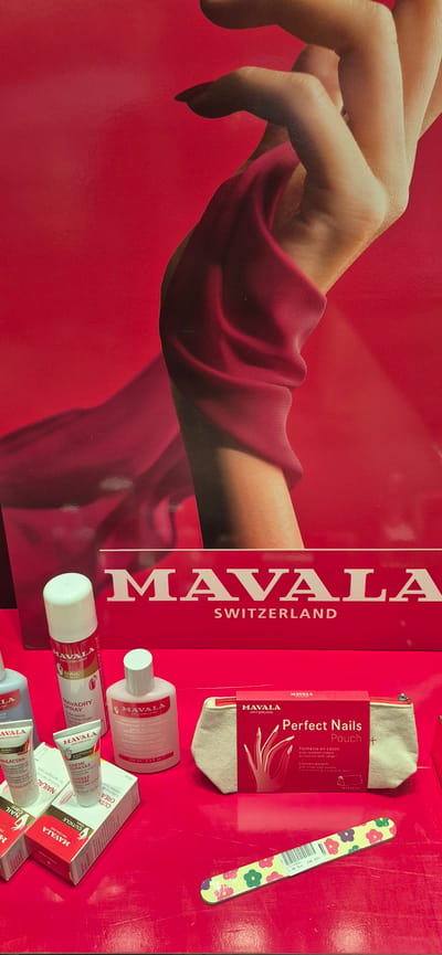 A red background features a close-up of a woman's hand, draped in red fabric. In the foreground, several Mavala nail care products are displayed on a bright red surface. These include bottles of nail polish remover and nail treatment sprays, as well as small tubes of cuticle cream. A red box labeled "Perfect Nails Pouch" is open to reveal a beige pouch and a nail file with a floral pattern. The scene appears to be a product display, possibly in a retail environment. The image does not contain any specific landmarks or details that would indicate a location in Póvoa de Varzim, Portugal.