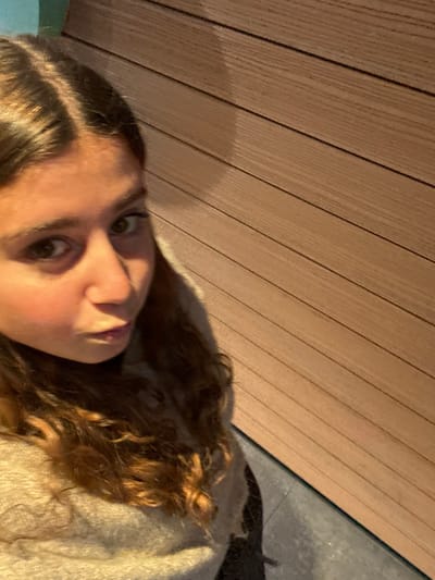 A close-up image depicts a young female partially framed on the left, looking directly into the camera from a slightly elevated angle. She has medium-brown, wavy hair parted down the middle. Her eyes are dark, and her lips are pursed. She wears a light-colored, possibly beige or cream-textured garment. The background consists of a wall clad with horizontal wooden panels of a light brown hue, displaying distinct parallel lines. A narrow section of dark grey or blue-grey flooring is visible at the bottom of the frame, adjacent to the wooden wall. The scene is indoors. The specific location within the city of Pombal, Portugal, cannot be determined from the visual evidence provided.