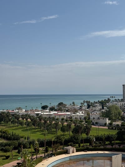 The image depicts a coastal scene in Erdemli, Turkey, featuring a clear blue sky with wispy clouds above the turquoise Mediterranean Sea. A row of white buildings lines the coastline, interspersed with palm trees and green vegetation. A green park area is visible, separating the buildings from a partially visible swimming pool. The overall scene portrays a serene coastal environment.