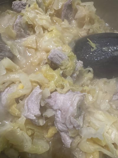 The image displays a close-up, top-down view of food actively cooking in a pot, located in Profesor Ishirkovo, Bulgaria. The contents consist of numerous irregular pieces of pale pinkish-white meat, interspersed with a substantial volume of light green and yellowish-white cabbage pieces, all submerged in a clear to slightly cloudy liquid. A dark, oblong utensil, consistent with a serving spoon or ladle, is partially visible on the right side, partially submerged in the food, with a small, light-colored food particle resting on its surface. No individuals are present.