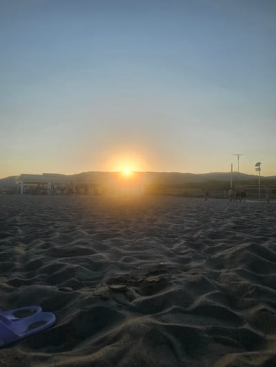 The image depicts a beach scene at sunset, likely in Castelldefels, Spain, as indicated by the context. The sun is low on the horizon, casting a warm, golden glow across the sky and the sand. The foreground is dominated by dark, rippled sand, suggesting the tide has recently receded or wind has sculpted its surface. In the mid-ground, there is a beach bar or restaurant with people seated at tables, some appearing to be enjoying the sunset. Further in the background, hills or mountains are visible, silhouetted against the bright sky. A tall pole with flags is also present, suggesting a recreational area or a landmark. The overall atmosphere is peaceful and serene, capturing the end of a day at the beach. There is a small, purple object, possibly a child's toy, visible in the lower right corner of the image. The weather appears to be clear, with no clouds obscuring the sunset. The time of day is clearly sunset.
