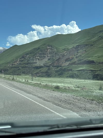 The image shows a landscape of a mountain range with a road running alongside it. The sky is blue with white clouds, and the mountains are covered in green vegetation. The road is gray and has a white line marking the lane. The image appears to have been taken from inside a vehicle, with a portion of the vehicle visible on the right side of the frame. Given the location information of Cholok, Kyrgyzstan, it is probable that this view is within the mountainous terrain of that region. There is no explicit indication of people, actions or interactions visible in the frame.