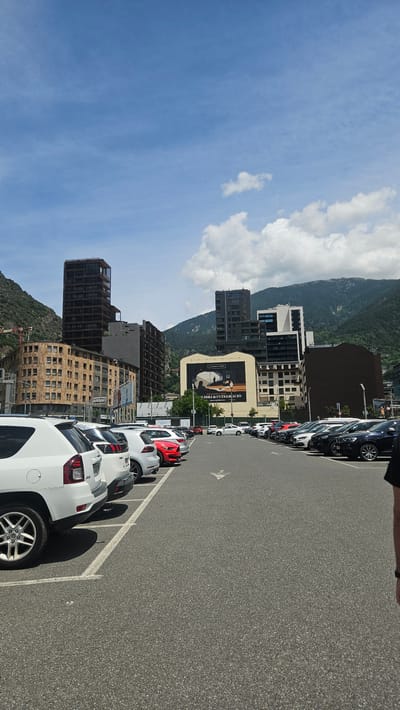 The image depicts a sunlit urban landscape within a mountainous valley, specifically in Andorra la Vella, Andorra, captured from a ground-level perspective within a large parking facility. The sky occupies the upper approximately one-third of the frame, displaying a clear blue expanse with scattered white cumulus clouds, more concentrated on the right side.
Mountains, predominantly green and rocky, rise steeply in the background on both the left and right, framing the city's built environment. On the far left, a construction crane is partially visible against the mountain.
The urban area comprises a mix of building types and ages. In the left-midground, a multi-story, light brown building with numerous windows stands next to a taller, dark brown modern skyscraper. Further right, a sequence of buildings includes a prominent cream-colored structure featuring a large billboard. The billboard displays a black and white image of two silhouetted figures in an indoor setting, with text in Catalan including "LLIBRI & INTEGRACIO" (Book & Integration). Behind and to the right of this building, several contemporary multi-story structures with varied facades (white and dark) are visible, extending towards the right side of the frame.
The foreground and mid-ground are dominated by an asphalt parking lot. White painted lines delineate individual parking spaces. Two main rows of parked automobiles are visible. The left row, closer to the camera, consists of at least five vehicles parked facing away from the viewer, including a white Jeep Compass SUV (partially visible rear-left), followed by another white SUV, a dark grey hatchback, and a red sports car. The right row contains numerous vehicles parked facing the viewer, including various SUVs and sedans in shades of white, black, grey, and red, extending into the distance. An arrow painted in white on the asphalt indicates vehicular traffic direction within the lot, pointing towards the buildings.
On the extreme lower-right edge of the frame, a partial view of a person's right arm and torso, clad in a dark short-sleeved garment, is visible. The scene suggests a bright, clear day in a bustling city integrated into a mountainous terrain.