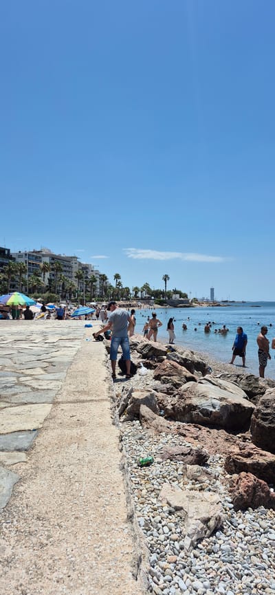 This is a full vertical shot of a beach scene in Paleo Faliro, Greece, on a clear, sunny day with a bright blue sky. The foreground features a concrete walkway bordering a rocky beach. A man in a grey t-shirt and blue denim shorts is standing on the walkway, facing away from the camera, holding a black bag. He appears to be interacting with something on the ground. Behind him, the rocky shore transitions to a sandy beach populated by numerous sunbathers and swimmers. Beach umbrellas in various colors dot the sandy area. In the background, several low-rise buildings, characteristic of a Mediterranean urban environment, are visible, interspersed with palm trees. Further out, the blue sea stretches to the horizon, where a tall skyscraper is faintly visible in the distance, along with more buildings and structures along the coastline. Several people are in the water, some wading and others swimming.
